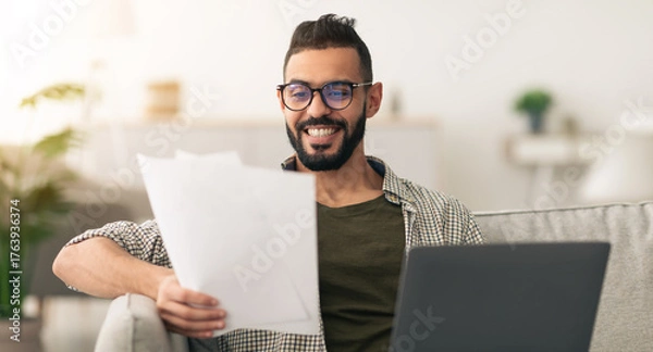 Fototapeta A man sits comfortably on a couch, smiling as he reviews printed documents while using a laptop. The cozy home office setting enhances the relaxed atmosphere.