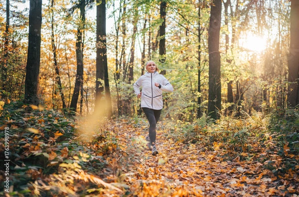 Fototapeta Middle aged woman jogging through autumn forest enjoying active lifestyle surrounded by colorful foliage. Morning fitness routine outdoors wellness, healthy lifestyle connection with nature concept