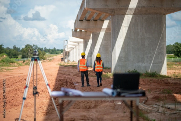 Fototapeta Engineers surveyor wearing helmets and reflective vests discuss construction blueprints work site surveying tripod and measurement equipment infrastructure or bridge construction industry.