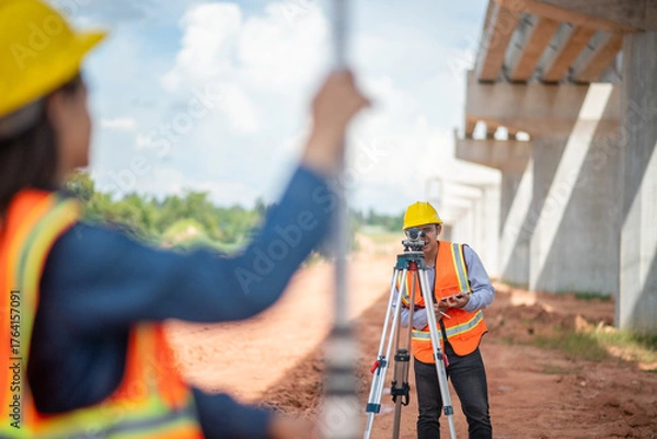 Fototapeta Engineers surveyor wearing helmets and reflective work site surveying tripod and measurement equipment infrastructure or bridge construction industry.