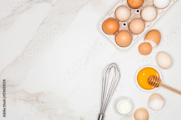 Fototapeta Baking cake ingredients. Milk, egg whites foam, egg beater, honey and eggshells on marble desk. Top view. Space for text.