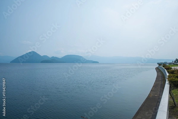 Fototapeta Calm lake with distant mountains and curved shoreline under clear sky at Lake Toya, Abuta, Hokkaido, Japan