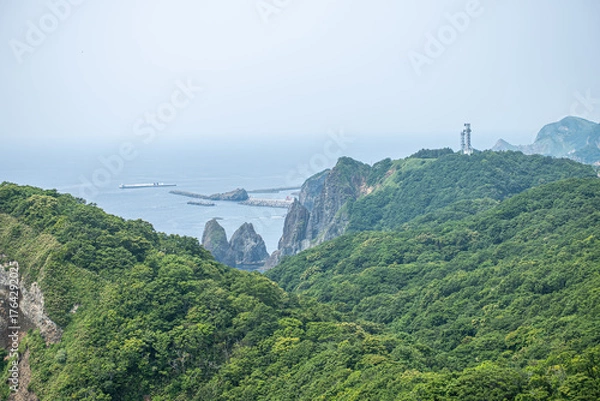 Fototapeta Lush green forested cliffs with lighthouse and ocean view on a hazy day at Cape Chikyu, Muroran, Hokkaido, Japan