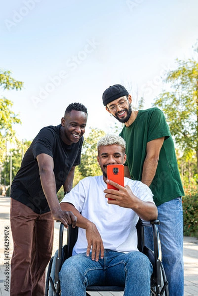 Obraz Vertical portrait of African American man in a wheelchair using phone together with male friends outdoors.