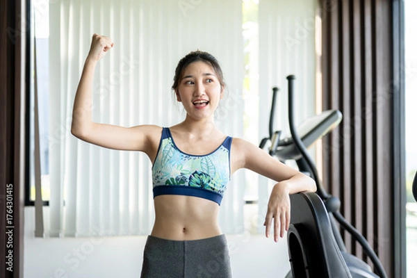 Fototapeta Pretty asian woman raising one hand making a fist standing aside exercise equipment in fitness gym.