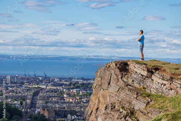 Fototapeta Scenic view of person hiker on cliff by city Edinburg and sea