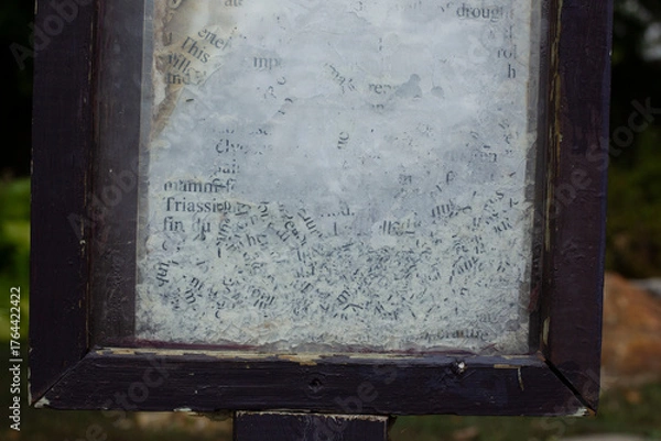 Fototapeta Close-up of weathered and partially erased text on a glass surface, framed by chipped dark wood. The faded black letters on a cloudy white background evoke age, decay, and forgotten information.