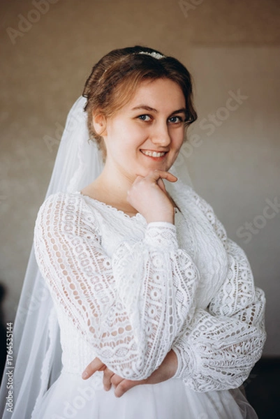 Fototapeta A beautiful bride in a white lace wedding dress smiles softly indoors. She stands by a window with sheer curtains, looking radiant and happy on her special day before the ceremony.