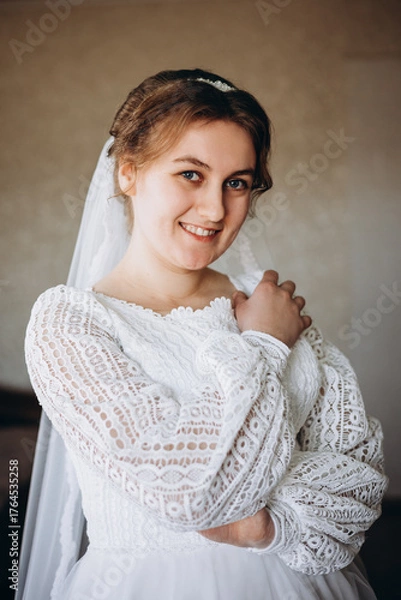 Fototapeta A beautiful bride in a white lace wedding dress smiles softly indoors. She stands by a window with sheer curtains, looking radiant and happy on her special day before the ceremony.