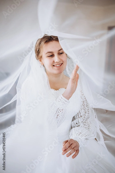 Obraz A smiling bride in a white lace wedding dress poses with her veil flowing gracefully around her. The soft fabric and natural light create a romantic and dreamy bridal portrait.