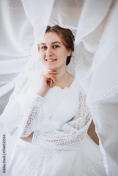 Fototapeta A smiling bride in a white lace wedding dress poses with her veil flowing gracefully around her. The soft fabric and natural light create a romantic and dreamy bridal portrait.