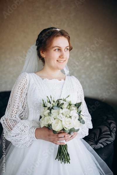 Fototapeta A beautiful bride in a white lace wedding dress holds a bouquet of white roses and greenery. She smiles softly, posing indoors before the wedding ceremony, looking elegant and serene.