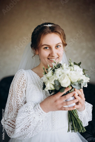 Fototapeta A beautiful bride in a white lace wedding dress holds a bouquet of white roses and greenery. She smiles softly, posing indoors before the wedding ceremony, looking elegant and serene.