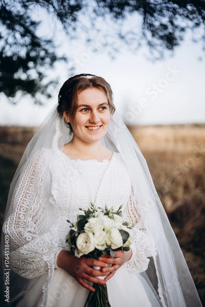 Fototapeta A bride in a beautiful white lace dress stands outdoors holding a bouquet of white roses. A calm and tender atmosphere of a wedding day in nature.