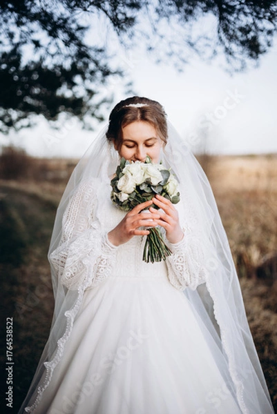Obraz A bride in a beautiful white lace dress stands outdoors holding a bouquet of white roses. A calm and tender atmosphere of a wedding day in nature.