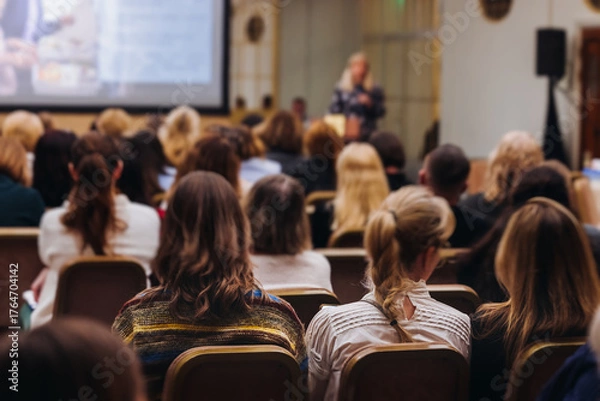 Fototapeta Female audience at the symposyum meeting, participants attendees in conference room hall listens to lecturer, group of women on a medical congress together listen to speaker on a stage at master-class