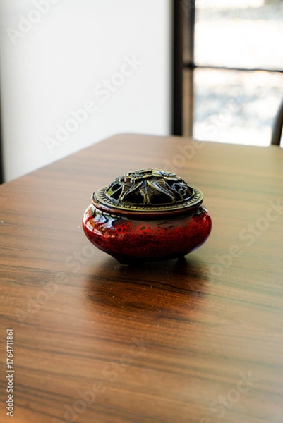 Fototapeta Elegant red ceramic incense burner with a detailed brass lid on a warm, polished wooden table. Perfect for relaxation, meditation, and decor