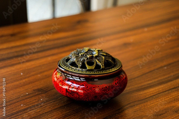 Obraz Elegant red ceramic incense burner with a detailed brass lid on a warm, polished wooden table. Perfect for relaxation, meditation, and decor