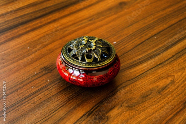 Obraz Elegant red ceramic incense burner with a detailed brass lid on a warm, polished wooden table. Perfect for relaxation, meditation, and decor