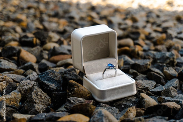 Obraz Stunning sapphire engagement ring in a white box placed on rugged dark gravel, symbolizing a bold, outdoor proposal or unique commitment