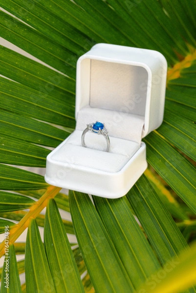 Obraz Exquisite blue sapphire and diamond engagement ring nestled in a white box on a vibrant green palm leaf, suggesting a romantic tropical proposal