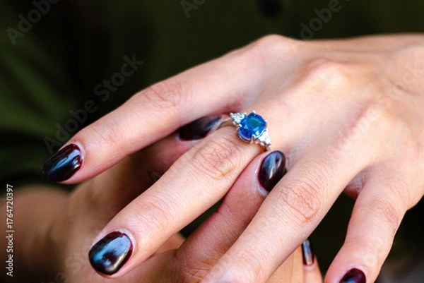 Fototapeta Close-up of a woman's hand wearing an elegant sapphire and diamond ring, with nails painted in a deep, rich burgundy color