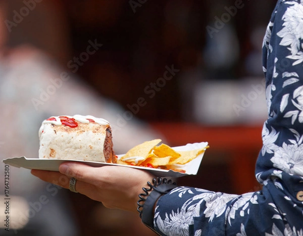 Fototapeta A person holding a paper tray with a burrito topped with sour cream and tomato salsa, served with tortilla chips at a street food market in Karlín, Prague.