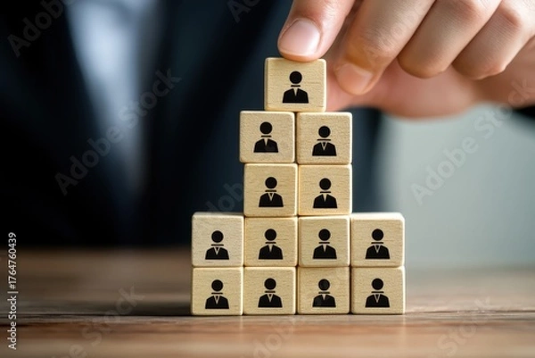 Obraz Building Success: A close-up view of a hand carefully placing a top cube adorned with a human icon atop a pyramid of similar cubes, symbolizing growth and unity