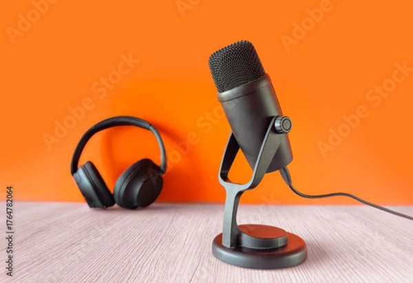 Obraz Black microphone and headphones on a light wooden table with an orange background, symbolizing podcasting or recording setup
