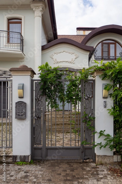 Obraz Ornate metal gate with climbing vines at entrance to luxury villa. Decorative security structure with stone pillars and wall sconces. Real photo