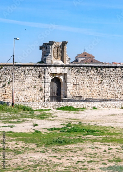 Obraz The medieval stone wall and entrance of the weathered Castillo de los Condes in Chinchón, Spain, framed by a lively field of yellow wildflowers and surrounded by a dirt road