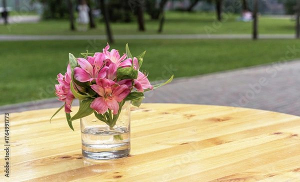 Obraz Delicate bouquet of pink lilies in glass vase on wooden table with blurred park background, romantic outdoor still life