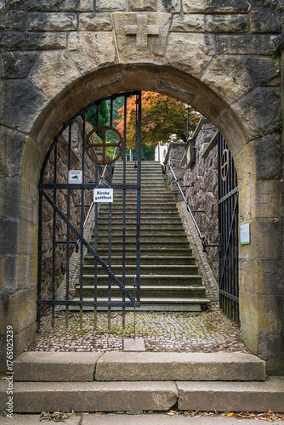 Fototapeta A stone gate with a cross symbol and an open iron grille bearing a "Church Open" sign leads to a steep staircase, the path to the church.