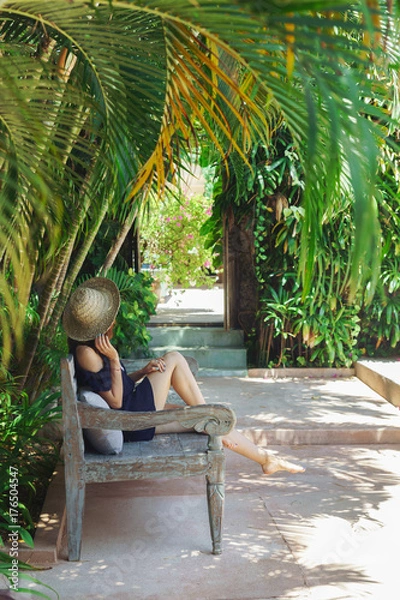 Fototapeta Young woman relaxing under the palm trees, hot sunny day,Bali.A young girl in a straw hat resting in the shade of coconut palms on a carved beautiful bench.Luxury holidays.