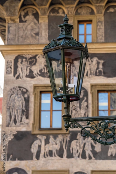 Fototapeta A vintage street lantern on an ornate bracket dominates the scene, set before a historic wall adorned with frescoes and arched windows, capturing urban heritag. Prague , Czech republic