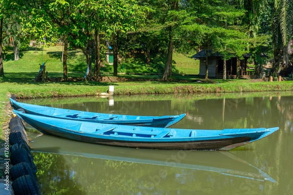 Fototapeta Two blue wooden boats are moored on the water's surface, with trees and a small wooden hut in the background. The scene is illuminated by bright sunlight, creating reflections on the water's surface.