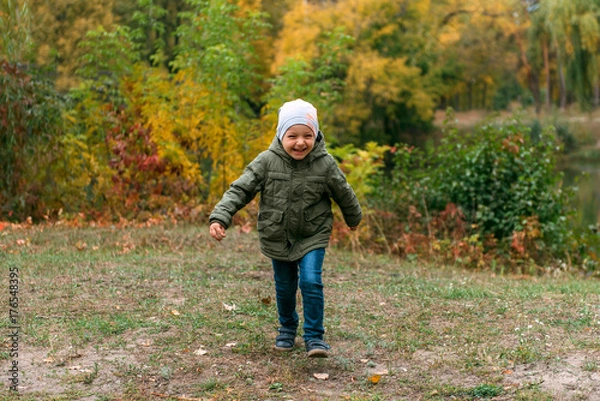 Fototapeta running boy child in a green jacket and a gray hat smiles against the background of the autumn forest of the park of the lake