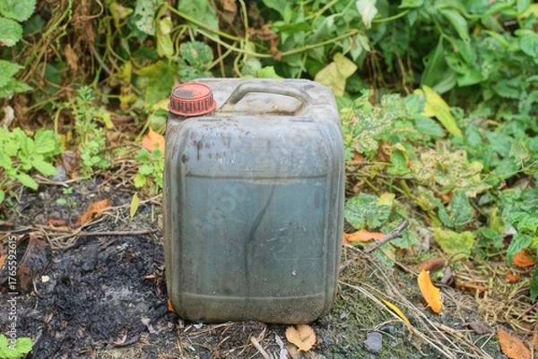 Obraz One old gray dirty plastic canister with a red cap filled with black liquid stands on the ground on a summer street