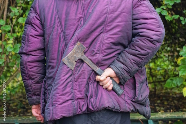 Fototapeta A man in a purple jacket holds a gray black axe in his hand behind his back on an autumn street