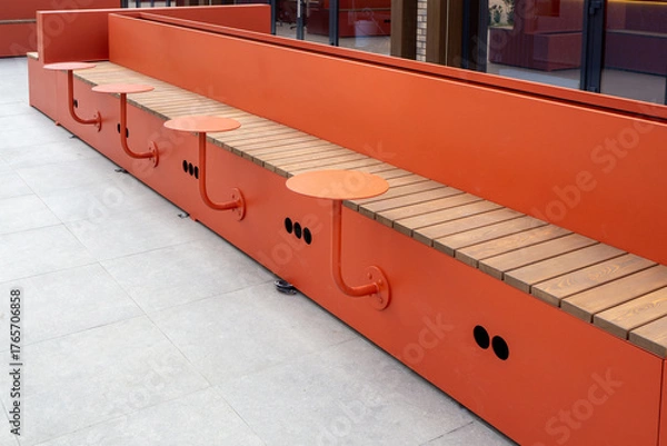 Obraz Long orange bench with integrated wooden seats and round tables on metal base at facade of modern building. Use for architecture, public spaces, urban furniture, landscape design. Real photo.