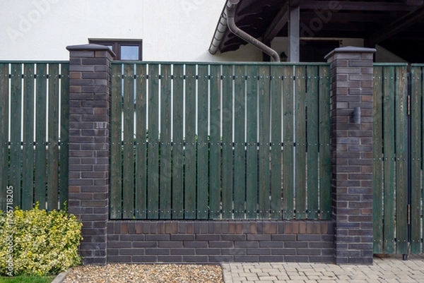 Obraz Dark green wooden fence with brick pillars and gravel base near residential house. For showcasing privacy fencing, property boundaries, and modern front yard entrance design. Real photo