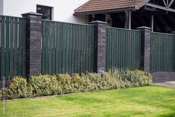 Obraz Green wooden fence with brick pillars and variegated hedge along neat lawn. For showcasing property borders, front yard landscaping, and low-maintenance garden edge design. Real photo.