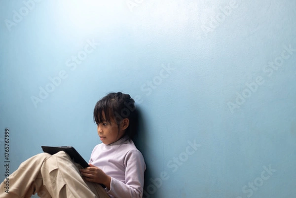 Obraz Asian little girl sitting and learning with digital tablet on blue wall background at home, concept of calm education, digital lifestyle, homeschooling, and smart learning technology.