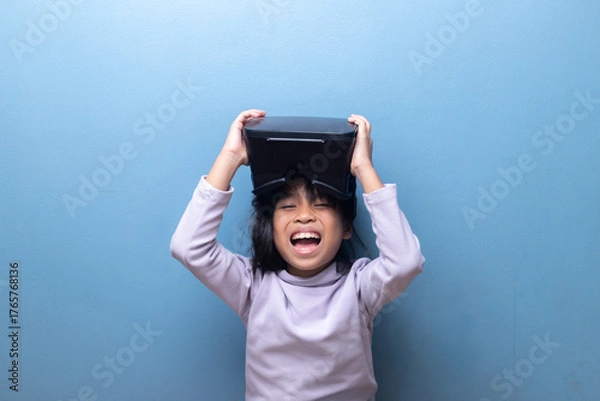 Obraz Happy Asian girl laughing with excitement while holding a VR headset. Child enjoying a fun virtual reality experience, metaverse gaming, or ed-tech on a blue background.