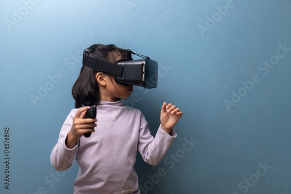 Obraz A young Asian girl wearing a virtual reality headset and holding a controller, exploring immersive digital experience, concept of future technology, education and metaverse.