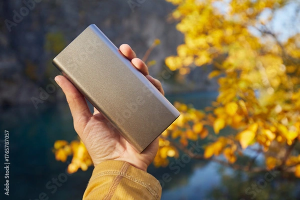 Fototapeta A man's hand holds a power bank with a charging phone against the backdrop of an autumn landscape