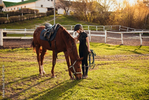 Fototapeta Young girl in equestrian gear enjoys a serene moment grazing a beautiful black horse in a green field, demonstrating the joy and connection between horse and rider.