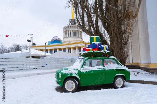 Fototapeta Small green retro car covered in snow with colorful wrapped presents on the roof, set against the backdrop of the main Kyiv VDNKh pavilion