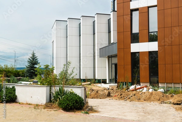 Fototapeta Contemporary office building under construction with contrast of white panels and brown wood-look facade panels, showing rough landscaping and dirt piles