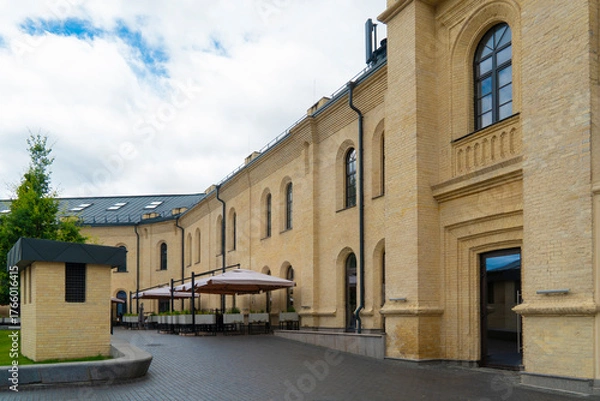Fototapeta Charming outdoor cafe terrace with light-colored umbrellas against the restored facade of a historic yellow-brick building on a cloudy summer day